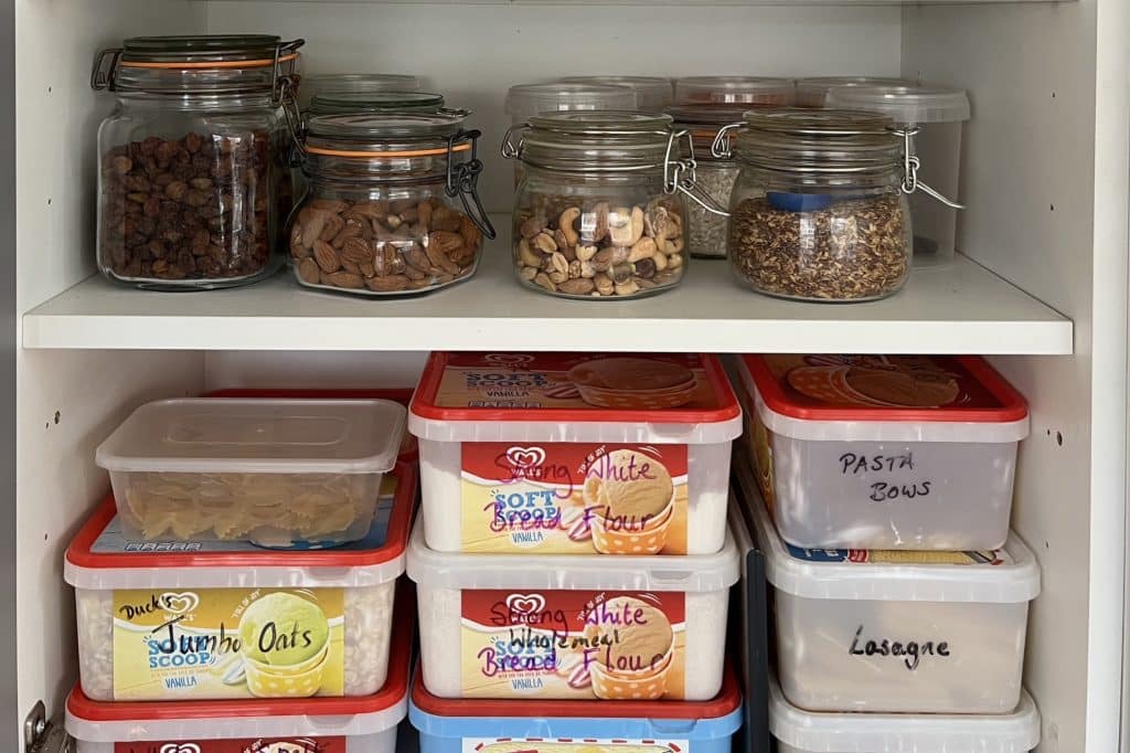 Our food cupboard with two shelves, one of glass containers full of seeds, nuts, sultanas rice and the other with plastic containers full of pasta, oats and flour.